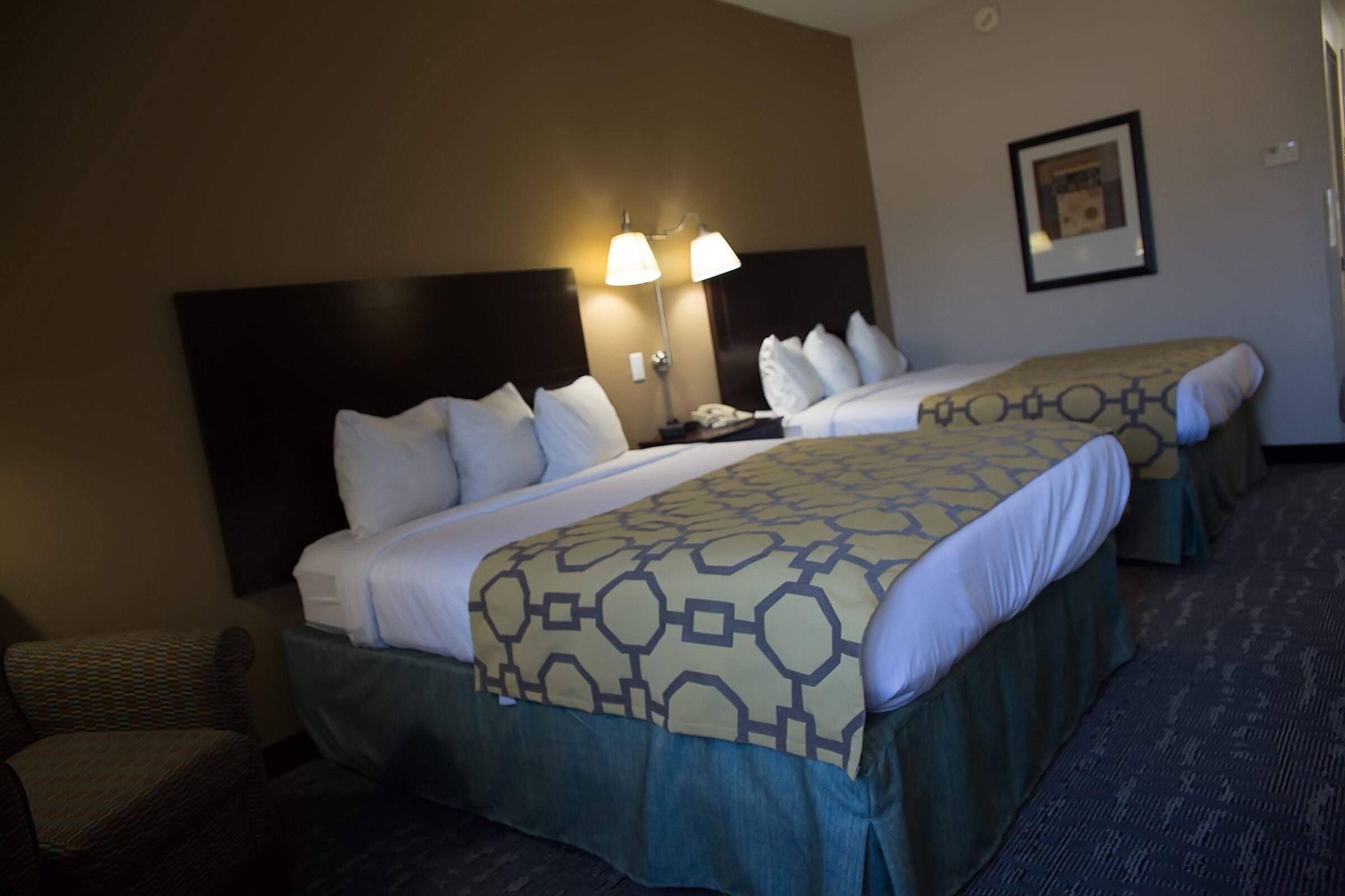 A hotel room with two beds, a headboard, a nightstand with a lamp, a chair, and a framed picture on the wall.