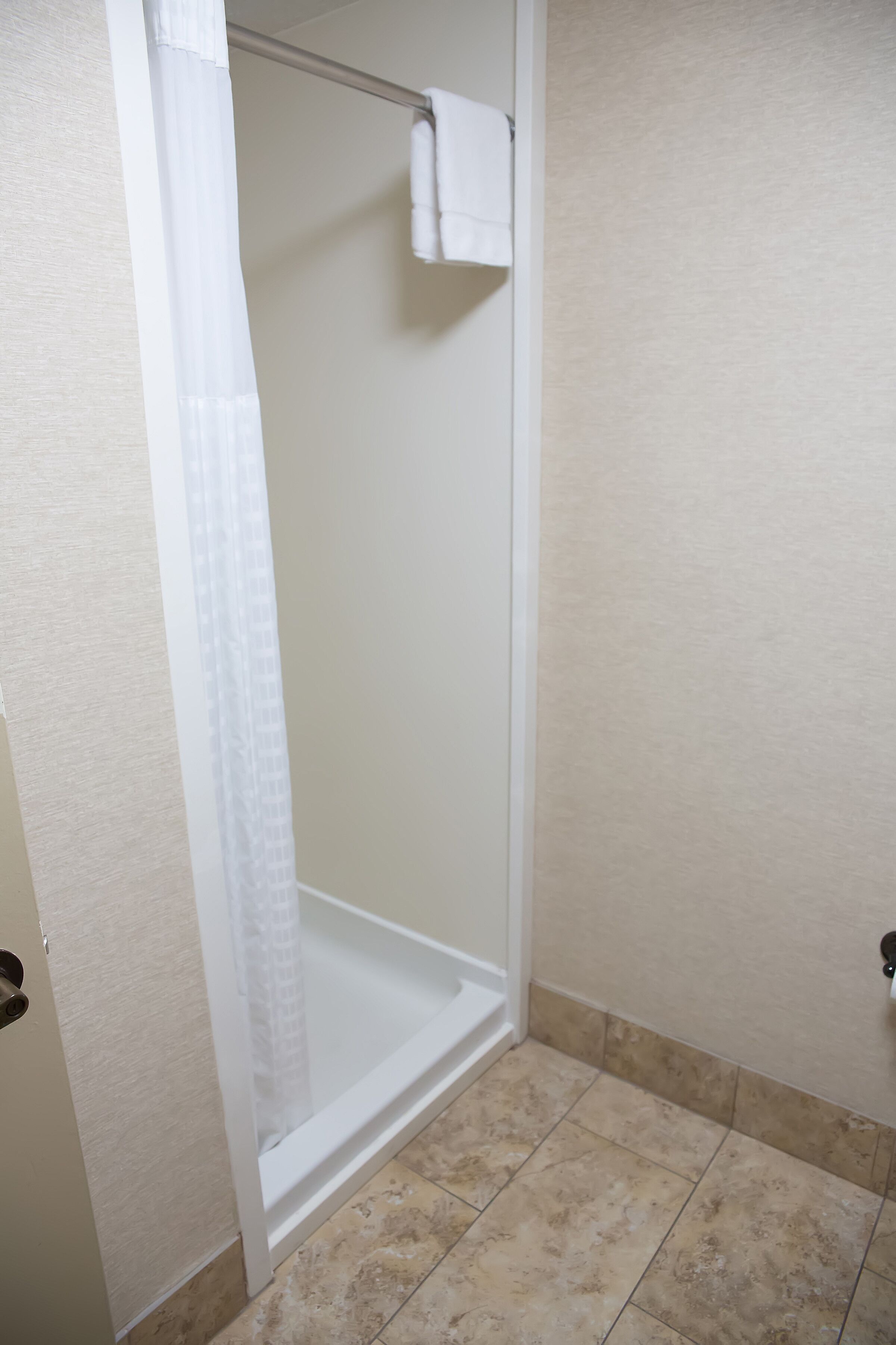 A bathroom with a glass shower enclosure, a white towel hanging on the shower rod, and beige tiled flooring.