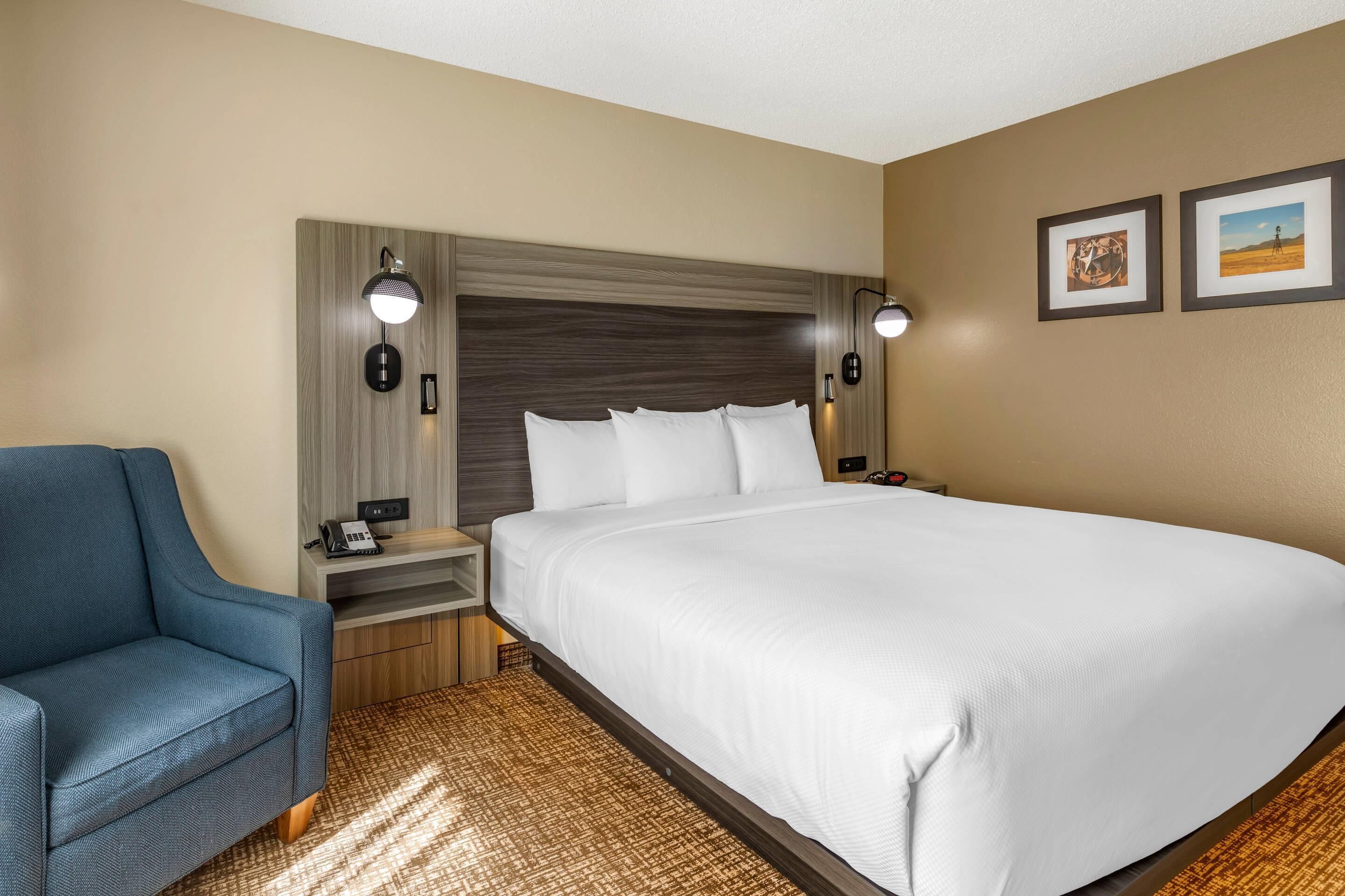 Une chambre d’hôtel avec un grand lit, un fauteuil bleu, une table de chevet en bois avec une lampe, et deux tableaux encadrés au mur.