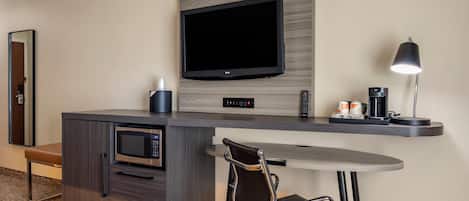 A hotel room with a flat-screen TV mounted on a wooden panel, a coffee maker, a microwave, a small table, and a chair.