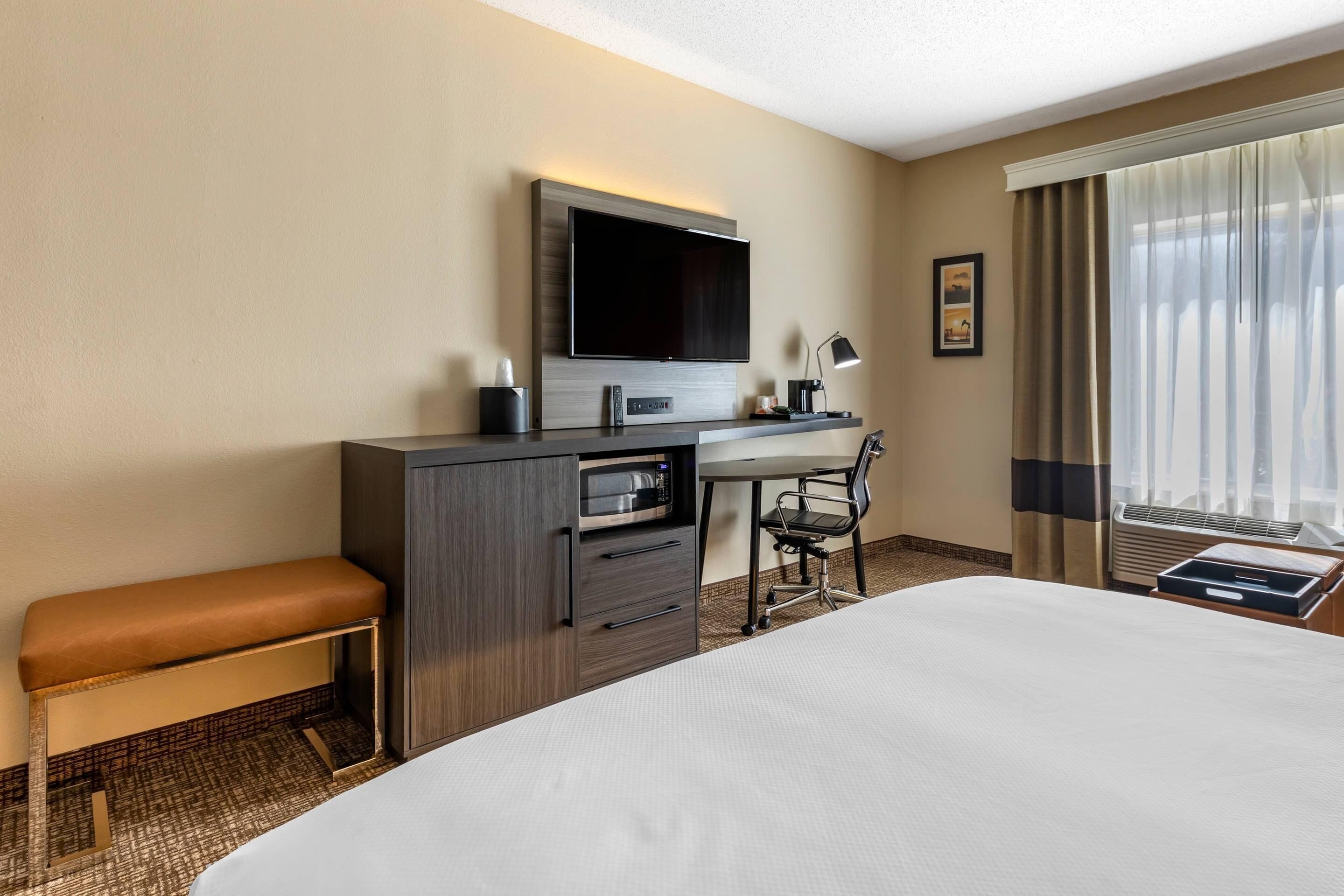 Habitación de hotel con una cama grande, un escritorio con televisor de pantalla plana, microondas, cafetera, una silla y una ventana con cortinas.
