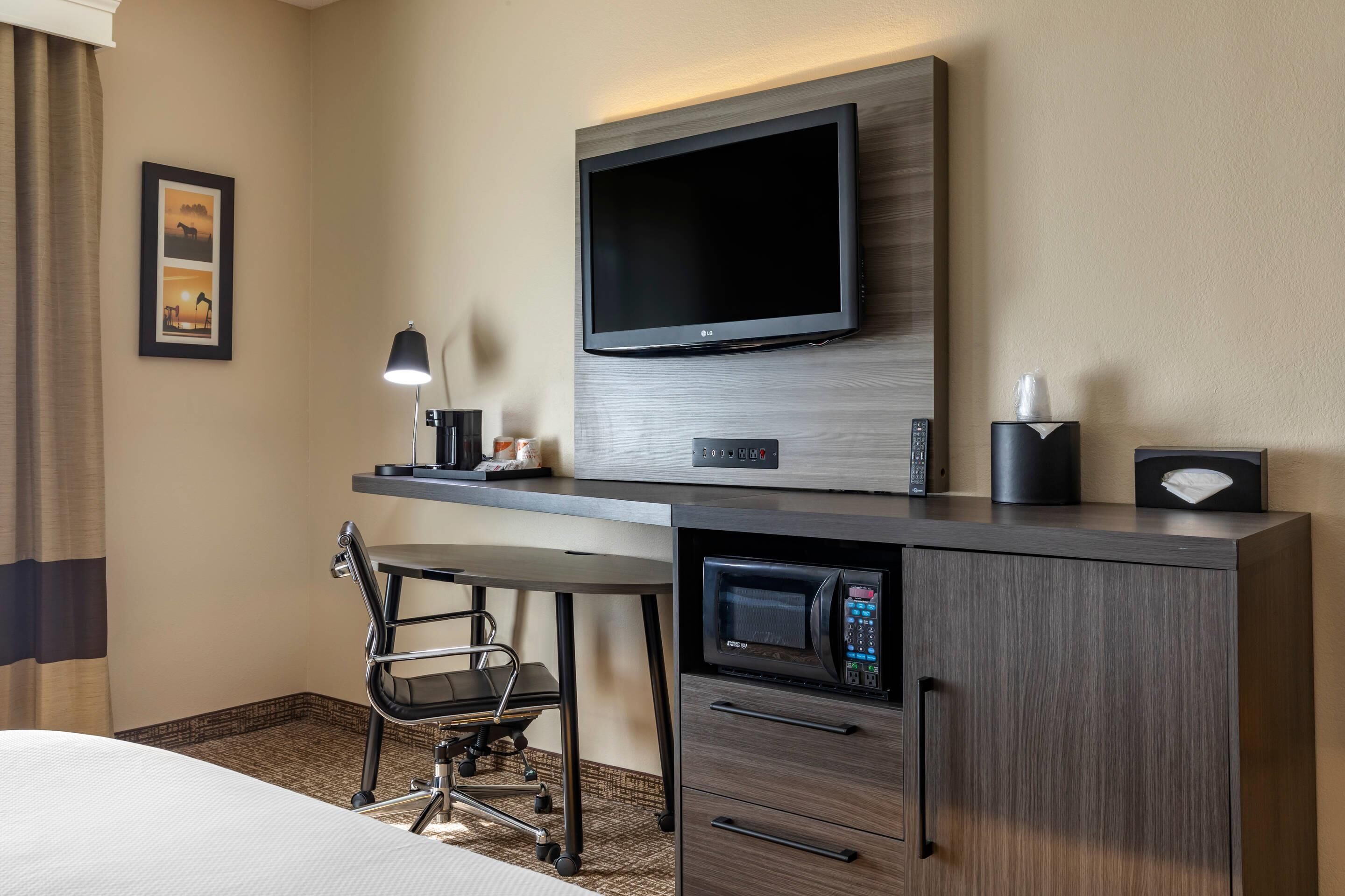 A hotel room with a bed, a desk with a chair, a flat-screen TV, a microwave, a remote control, and a framed picture on the wall.