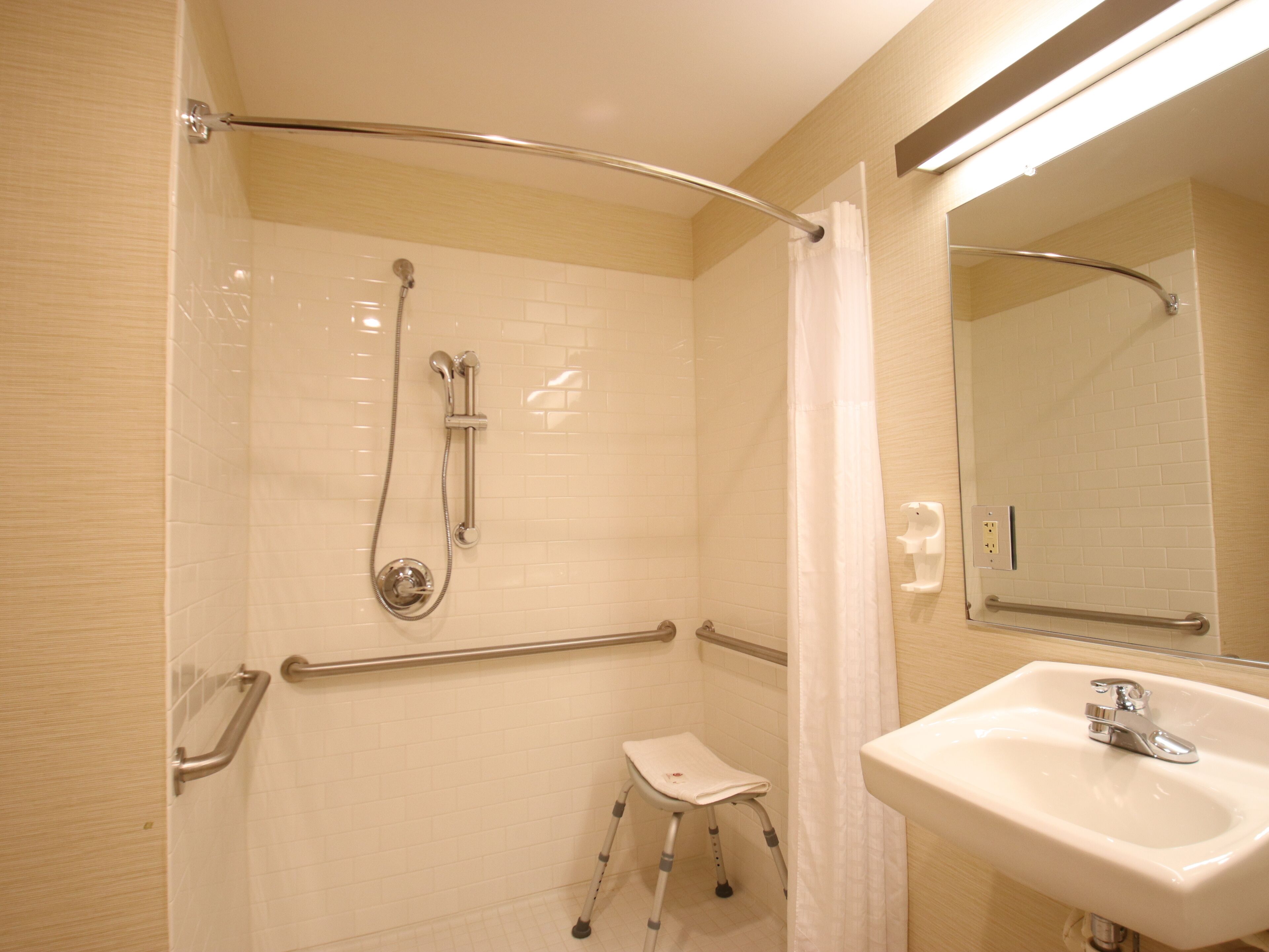 A bathroom with a walk-in shower, grab bars, a small stool, and a sink with a mirror.