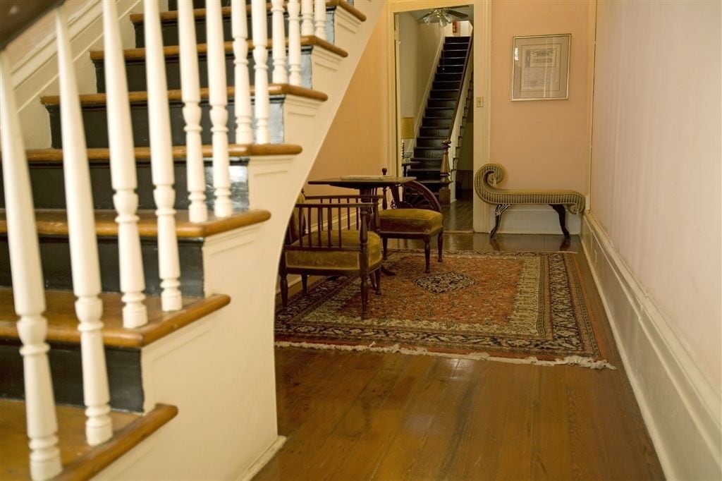 Una escalera con balaustres blancos y escalones de madera que conduce a un rellano. En el rellano hay una silla de madera tradicional y una mesita con un tapete.
