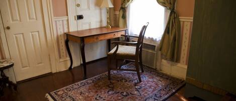 A room with a wooden desk, a chair, a patterned rug, a lamp, and a window with curtains.