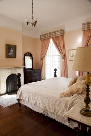 A bedroom with a large bed, a dresser, a mirror, a lamp, and a fireplace. - Fairchild House Bed & Breakfast (New Orleans)