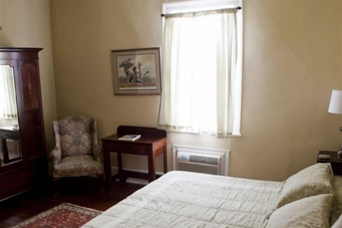 A bedroom with a bed, a chair, a table, a radiator, and a window with curtains.