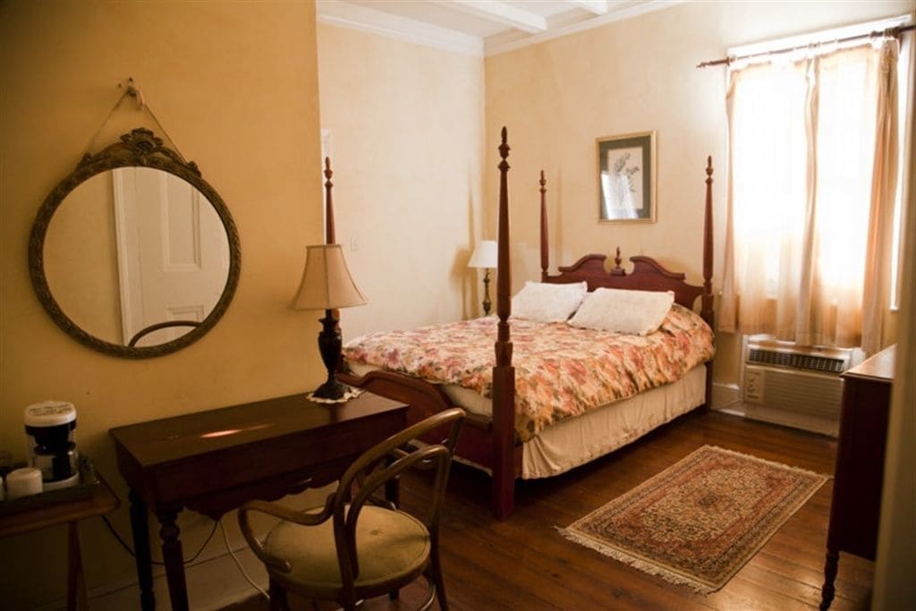 A bedroom with a four-poster bed, a wooden desk, a chair, a mirror, a bedside table, and a radiator.