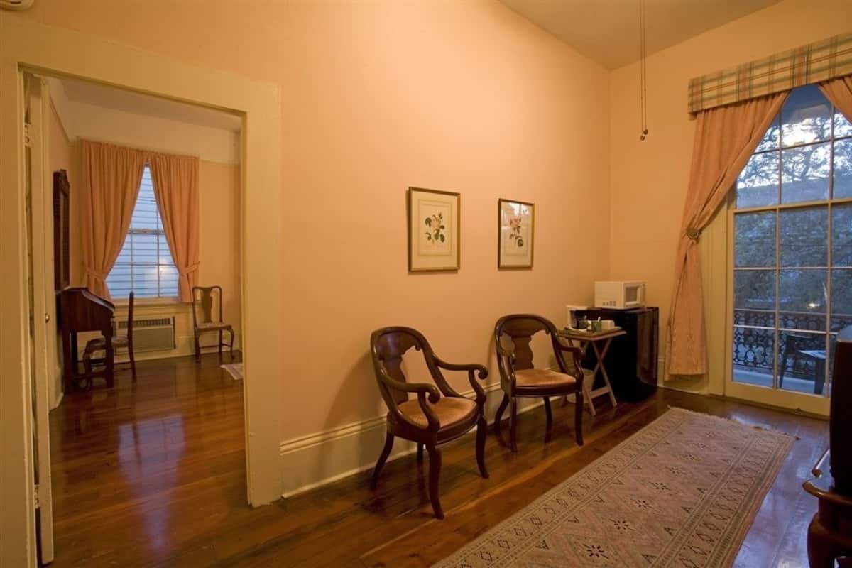 A room with wooden flooring, two framed pictures on the wall, a small table with a microwave, and a large window with curtains.