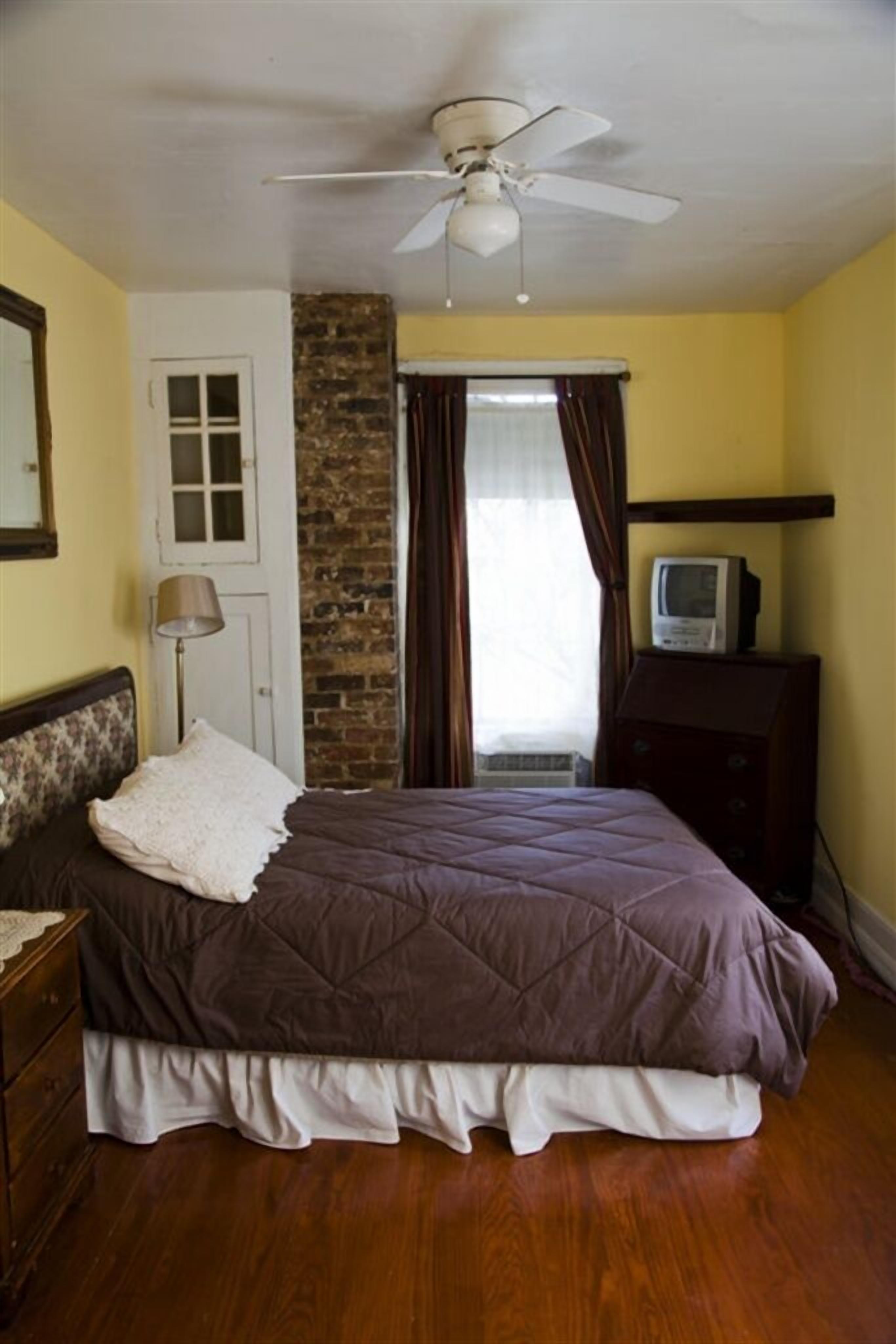 A bedroom with a bed, a ceiling fan, a television on a dresser, and a brick wall.