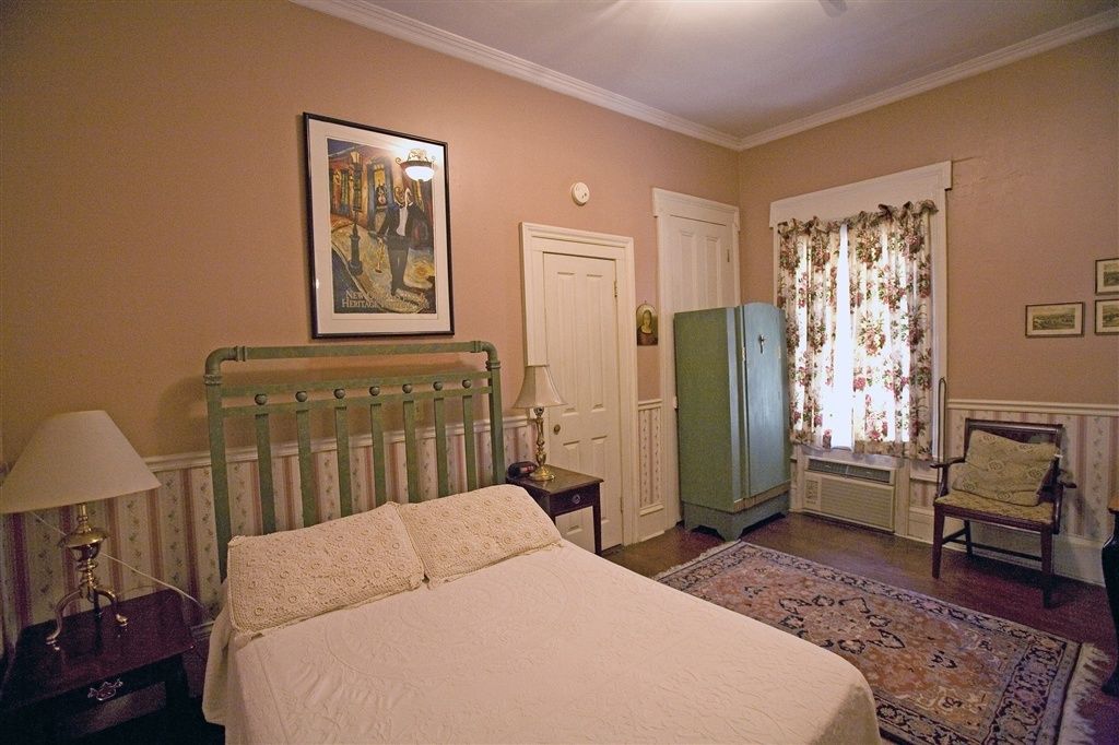 A bedroom with a bed, a nightstand, a lamp, a dresser, a chair, a window with curtains, and a framed picture on the wall.