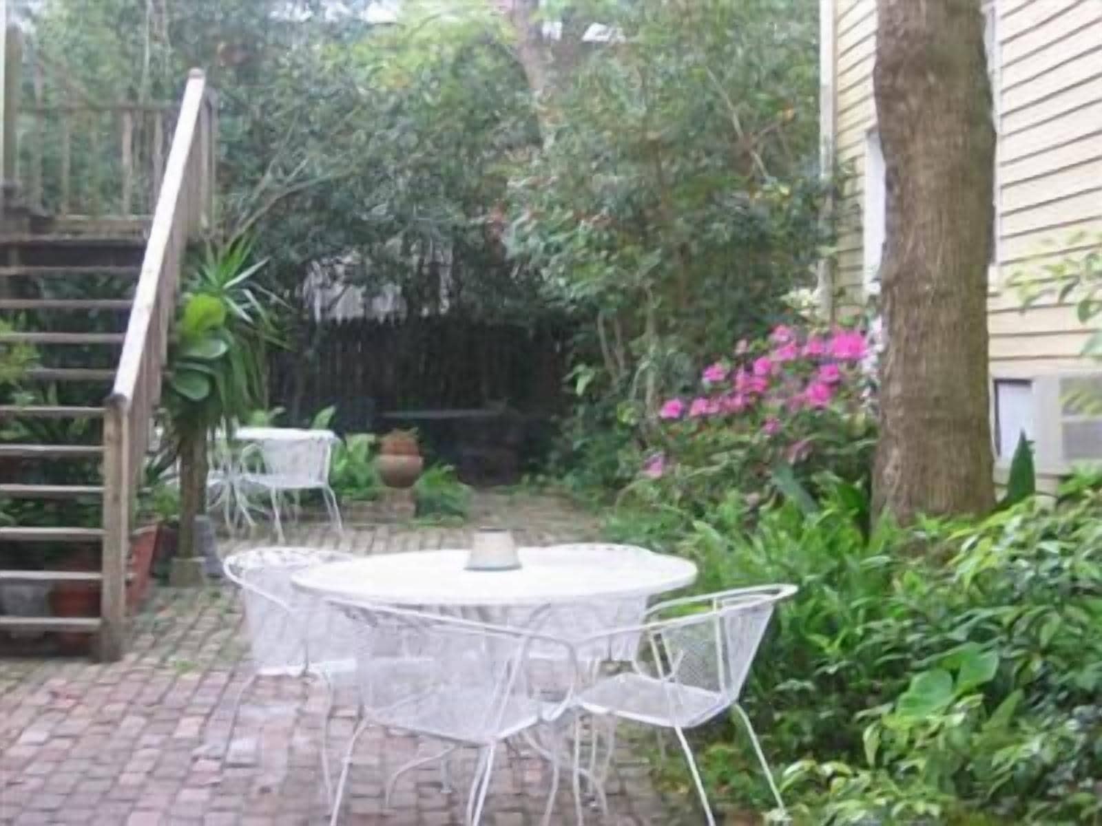 A patio with a round table and chairs, surrounded by greenery and a wooden staircase.