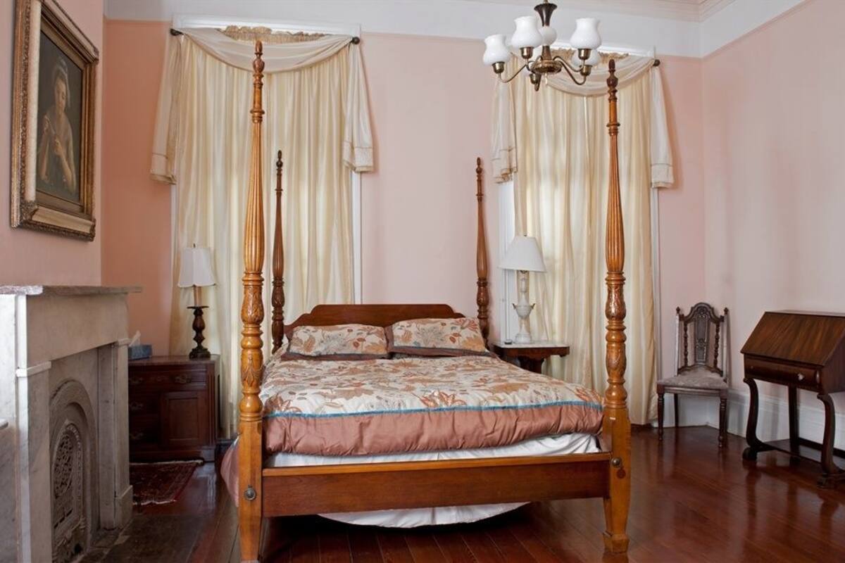 A four-poster bed with a floral bedspread, a wooden nightstand with a lamp, a chair, and a desk with a lamp.
