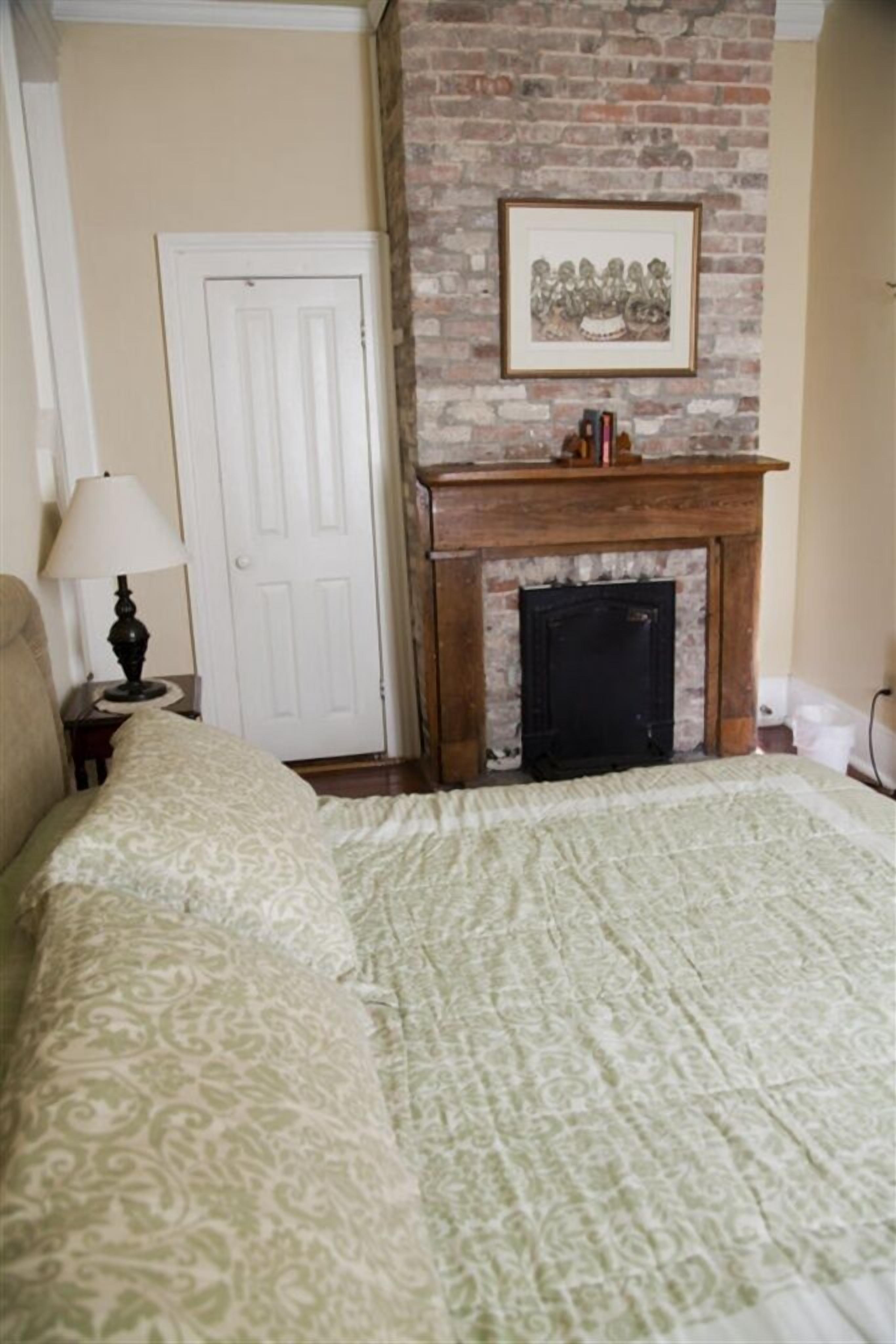 A bedroom with a brick fireplace, a bed with patterned bedding, a nightstand with a lamp, and a framed picture above the fireplace.