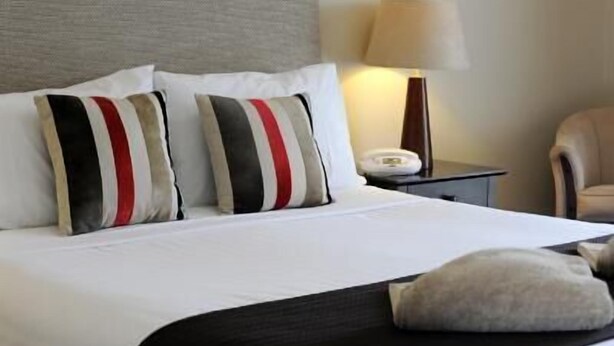 A neatly made bed with white bedding and striped pillows, a bedside lamp, and a chair in a hotel room. - Central City Motor Inn (Bakery Hill)