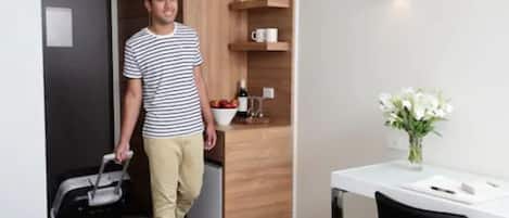 Habitación de hotel con armario de madera, pequeña cocina con un lavamanos y un frutero, mesa blanca con un florero, y una cama con una maleta.