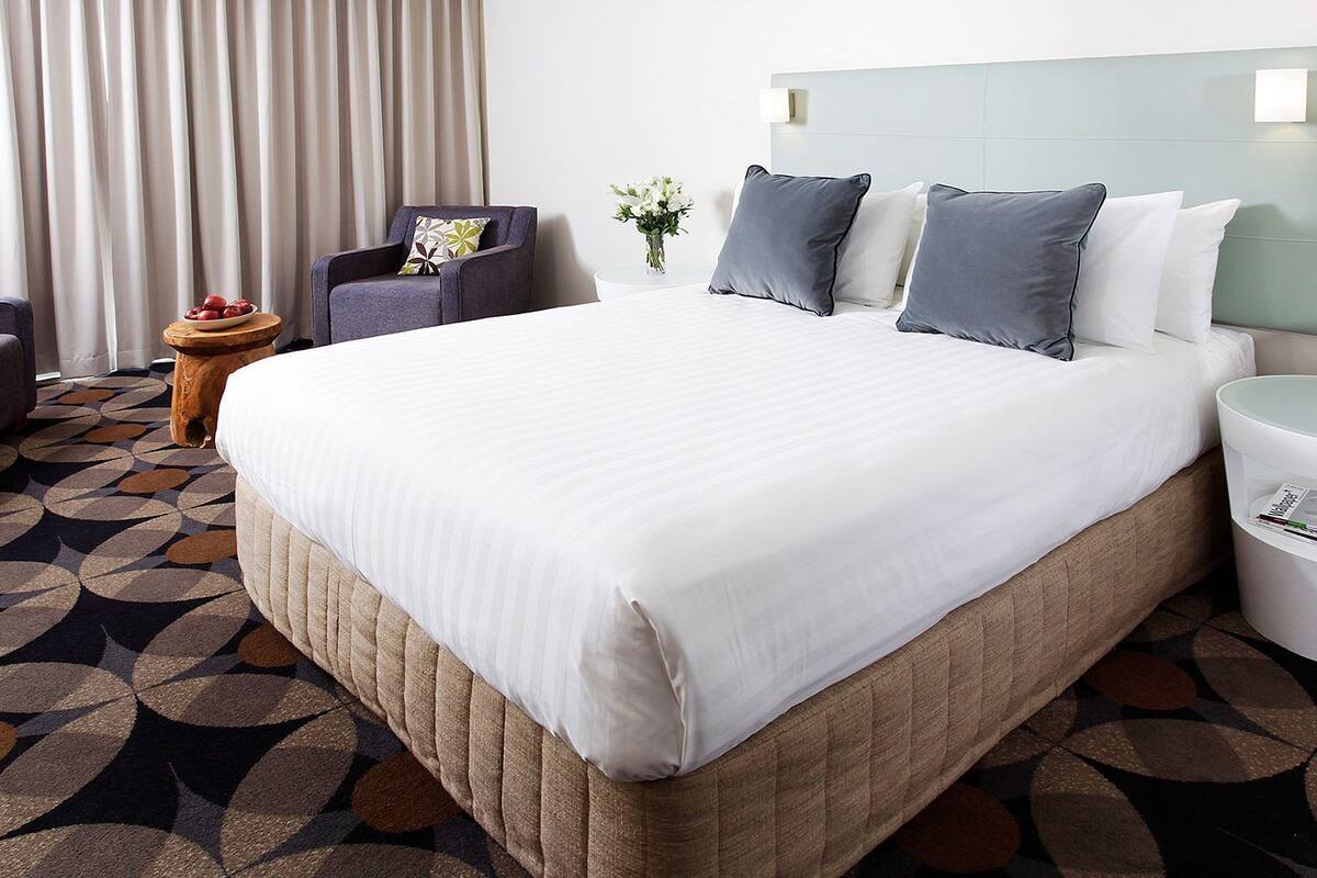 A hotel room with a large bed, a chair, a small table with a vase of flowers, and a trash bin.