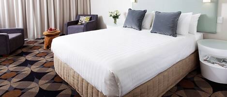 Habitación de hotel con una cama grande, una silla, una mesita con un florero y un cesto de basura.