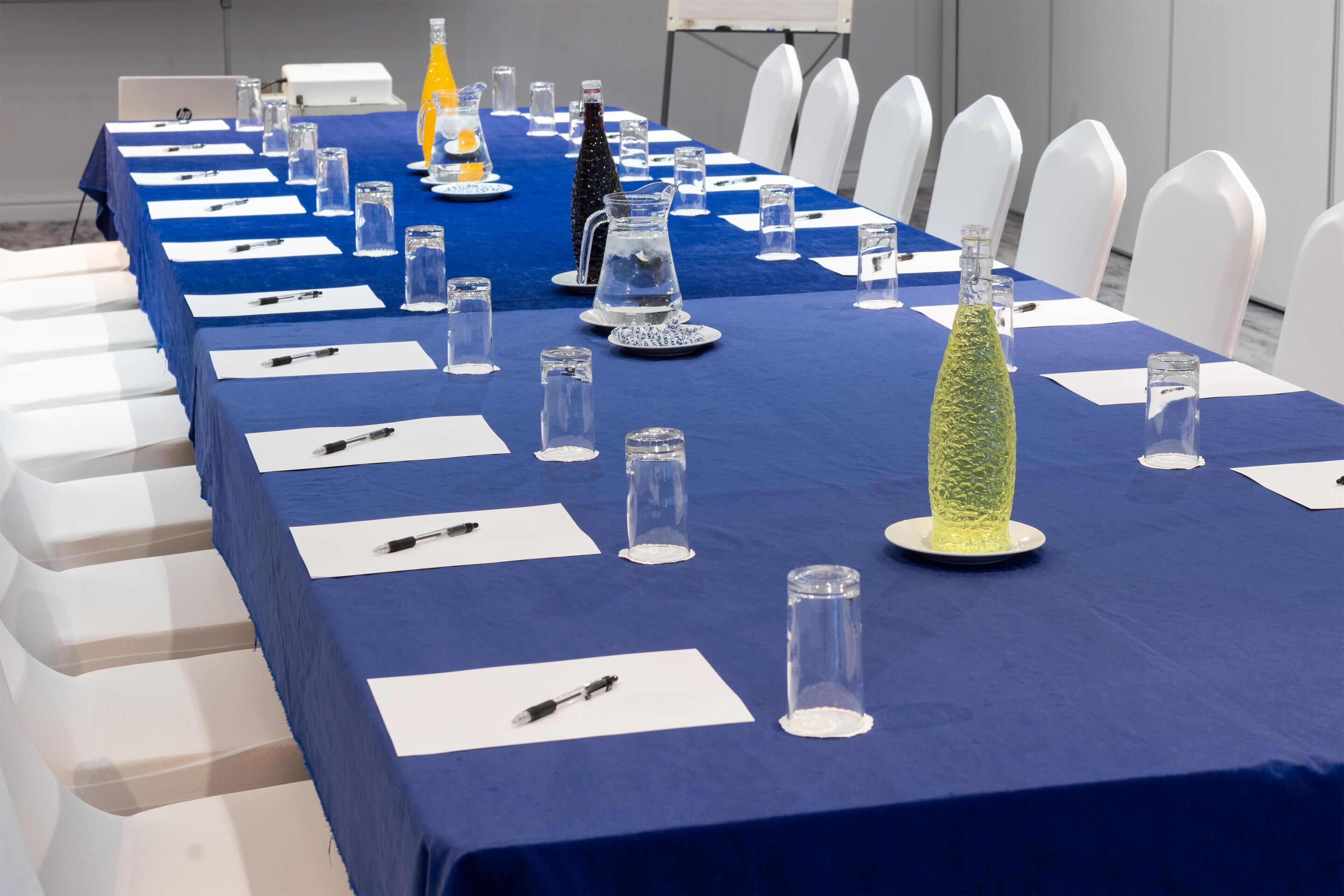 A conference room with a long table set for a meeting, white chairs, and a blue tablecloth.