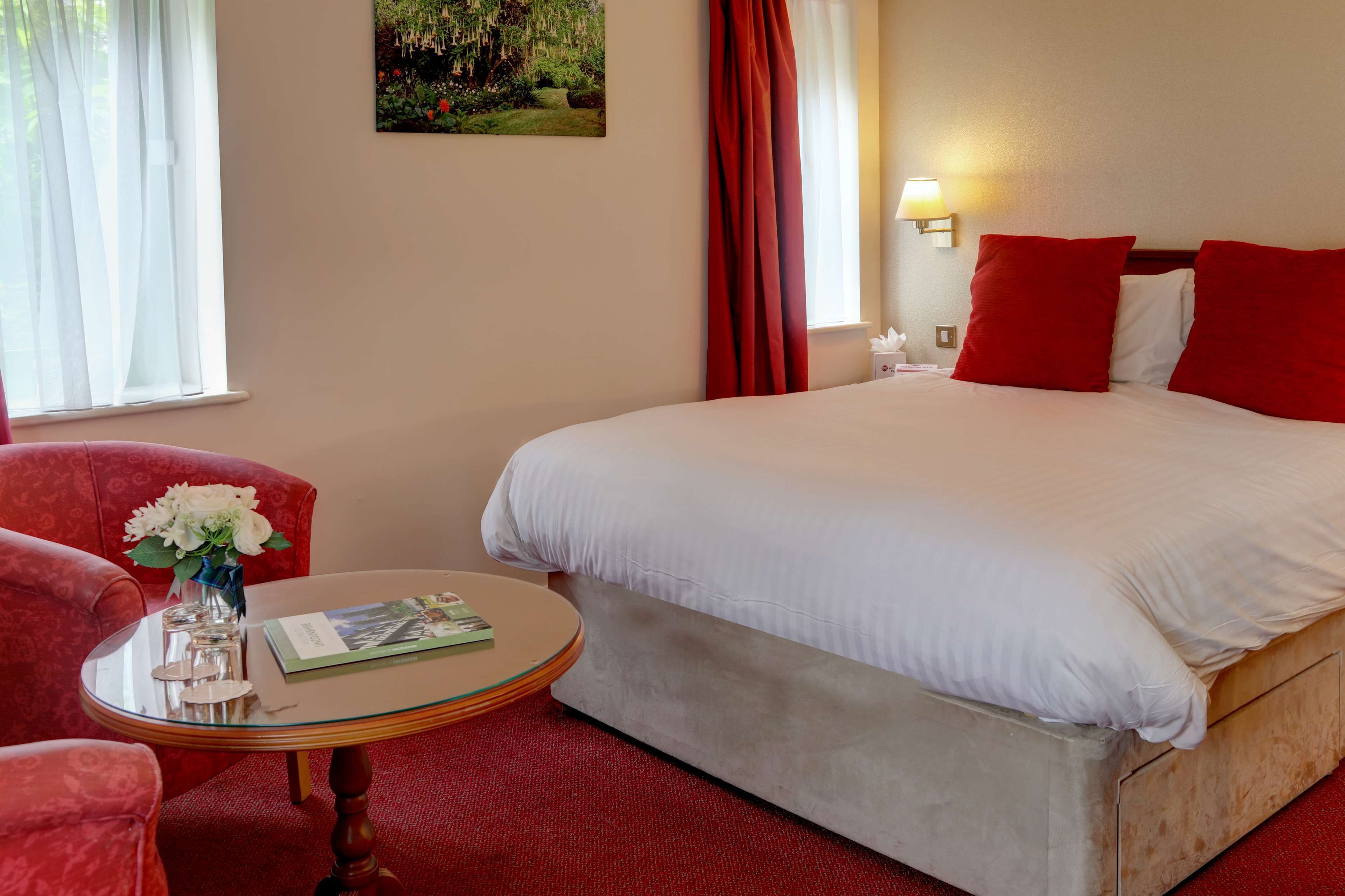 Una habitación de hotel con una cama grande, cortinas rojas, una mesita con un jarrón de flores y un cuadro en la pared.