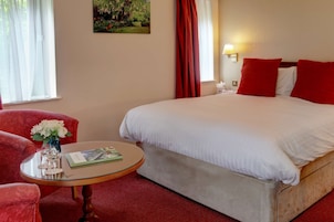 Una habitación de hotel con una cama grande, cortinas rojas, una mesita con un jarrón de flores y un cuadro en la pared.