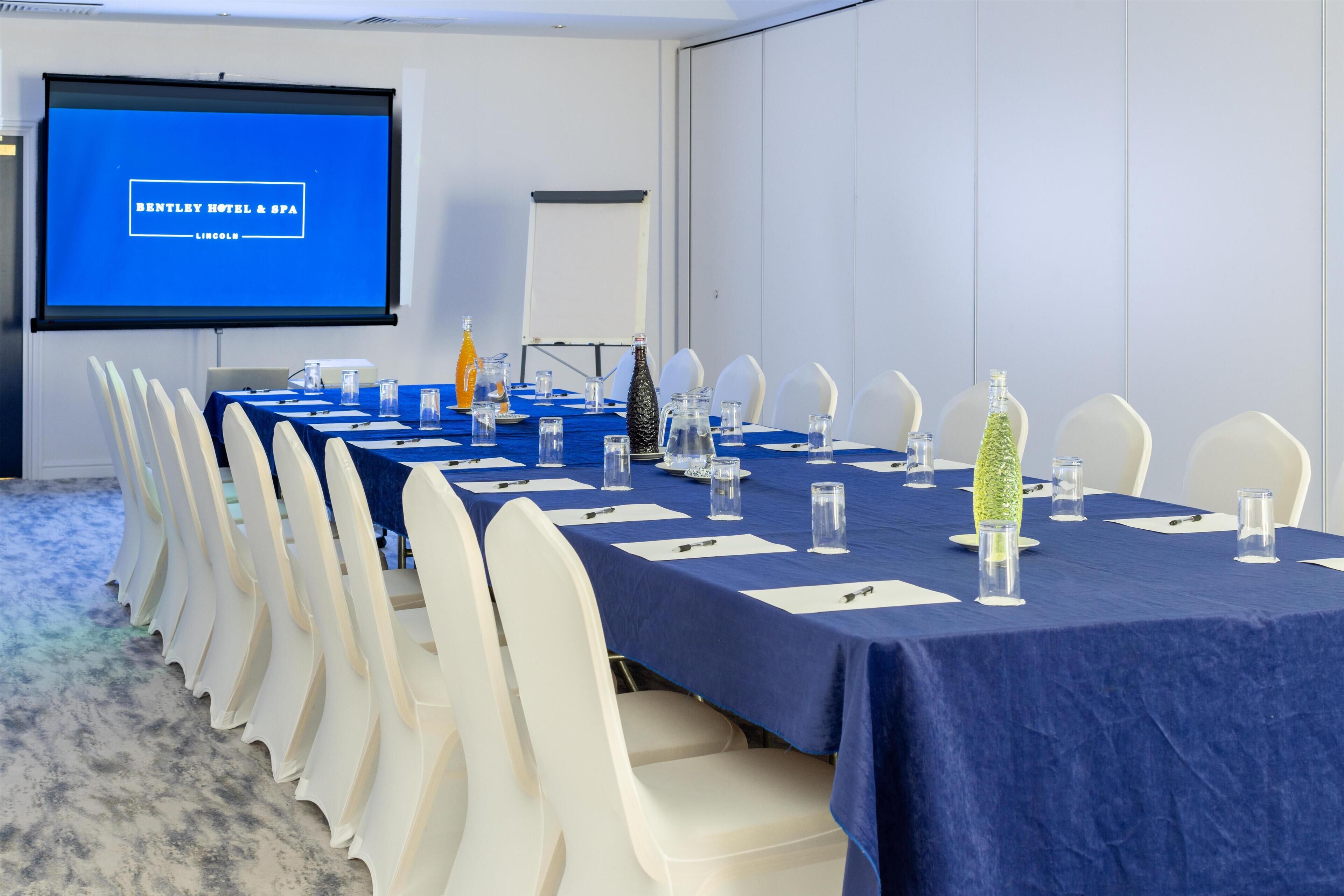 Un salón de conferencias con una larga mesa, sillas blancas, una pantalla de proyector y un mantel azul.