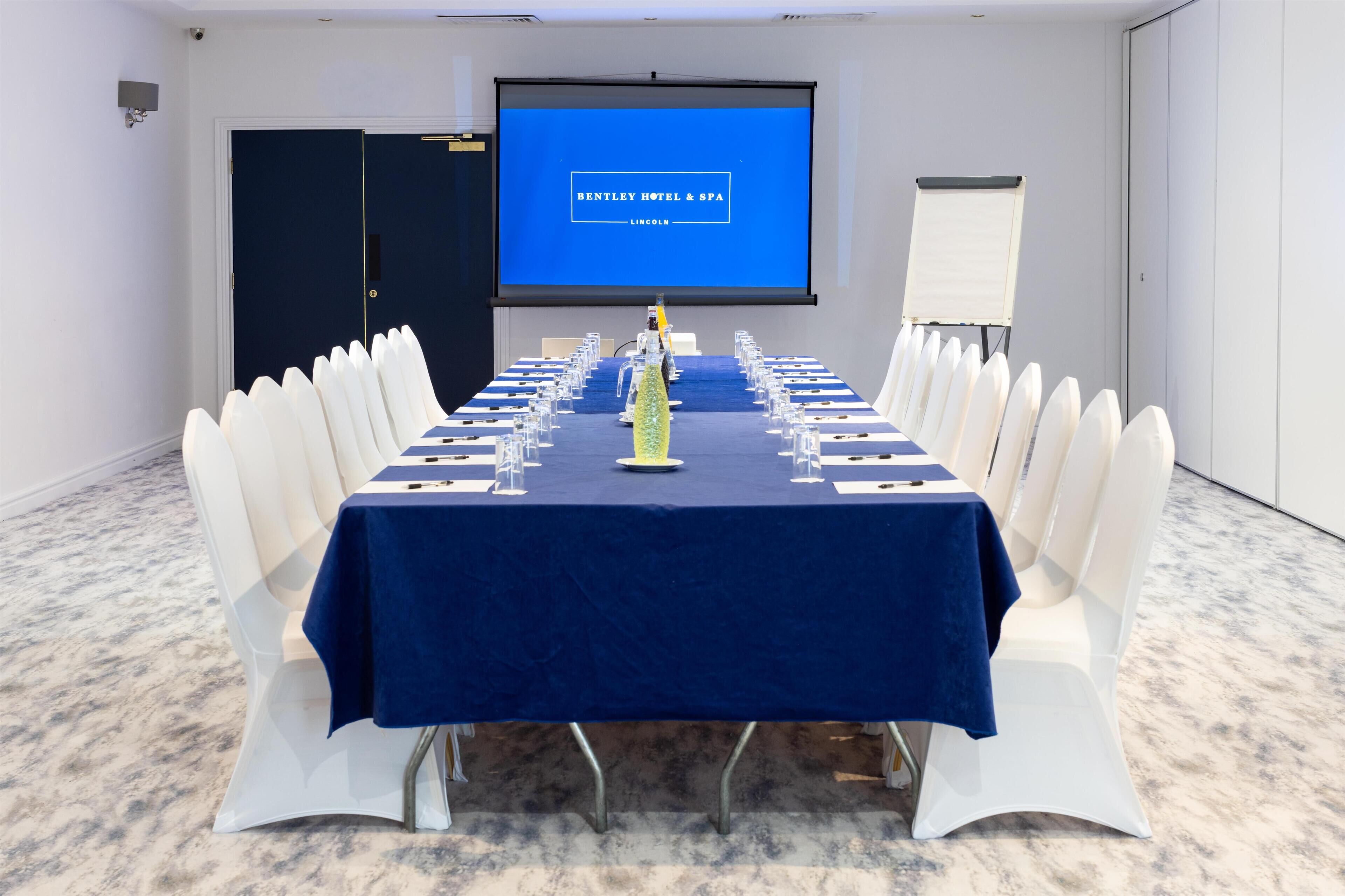 Een vergaderruimte met een lange tafel, witte stoelen, een projectorscherm waarop 'BENTLEY HOTEL & SPA LINCOLN' staat, en een whiteboard.