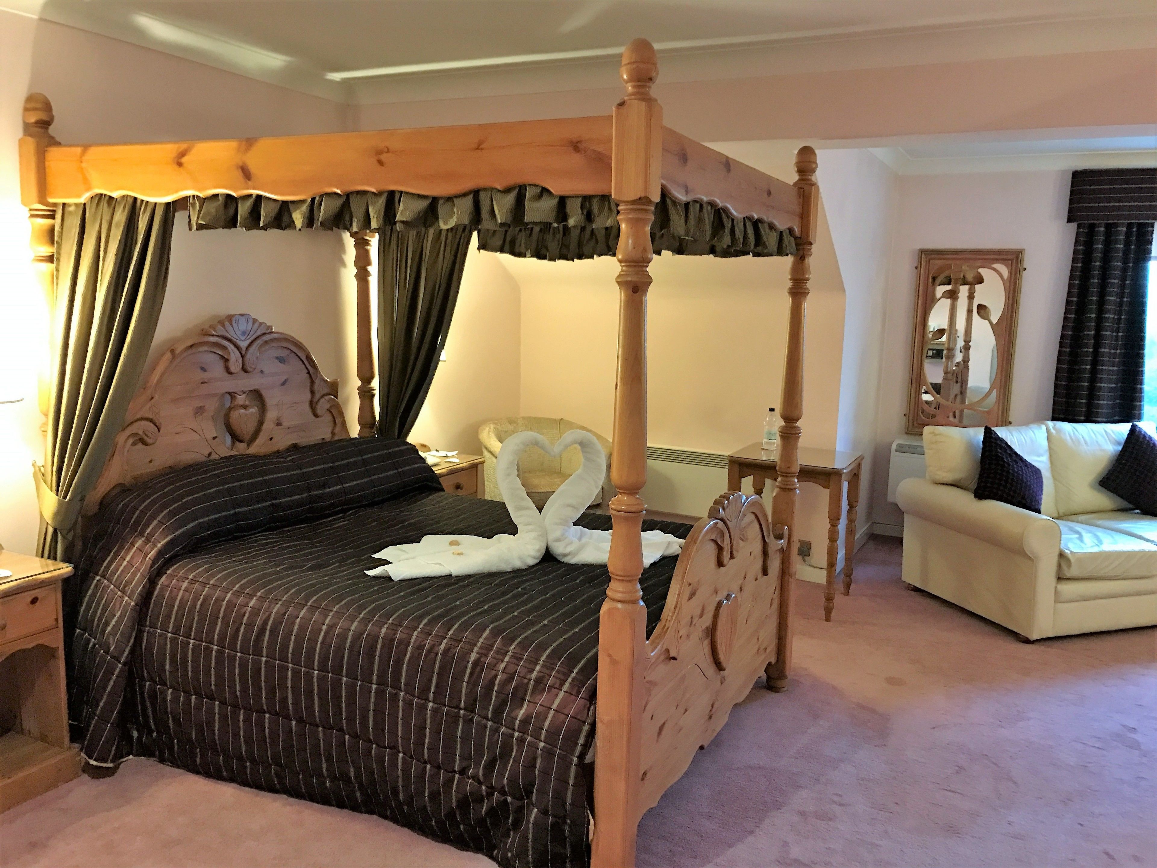 A four-poster bed with a heart-shaped pillow arrangement, a cream-colored sofa, and a mirror on the wall.