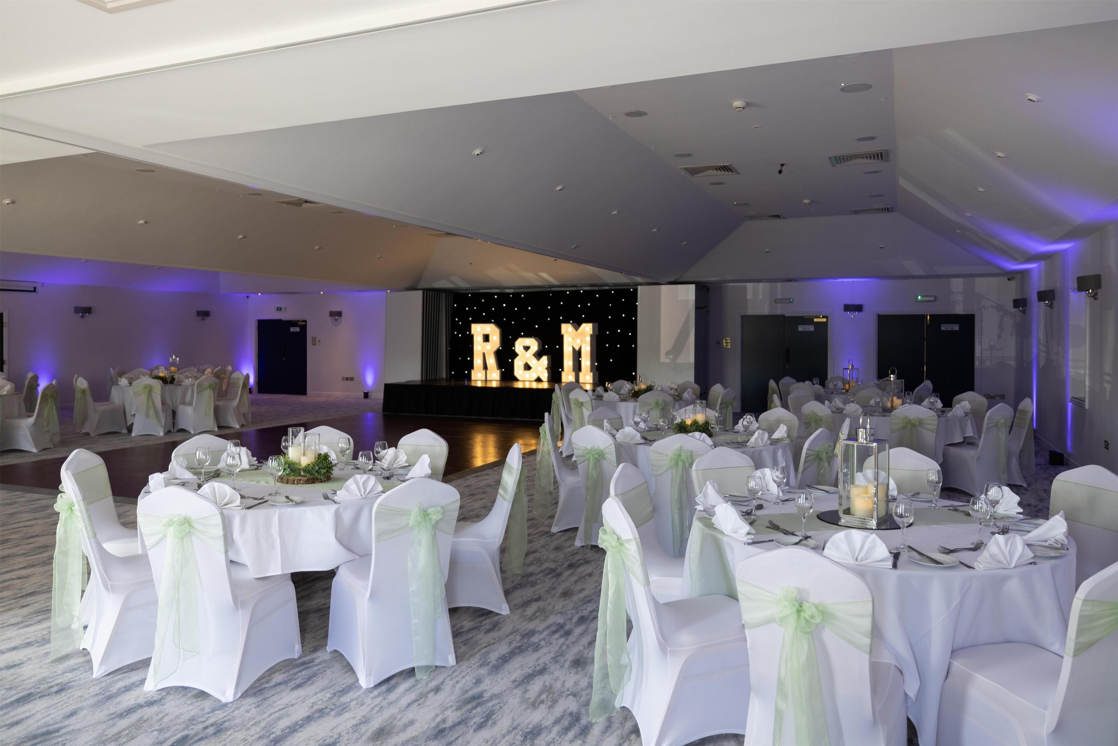 A banquet hall with round tables, white chairs with green sashes, and a stage with illuminated 'R&M' letters.