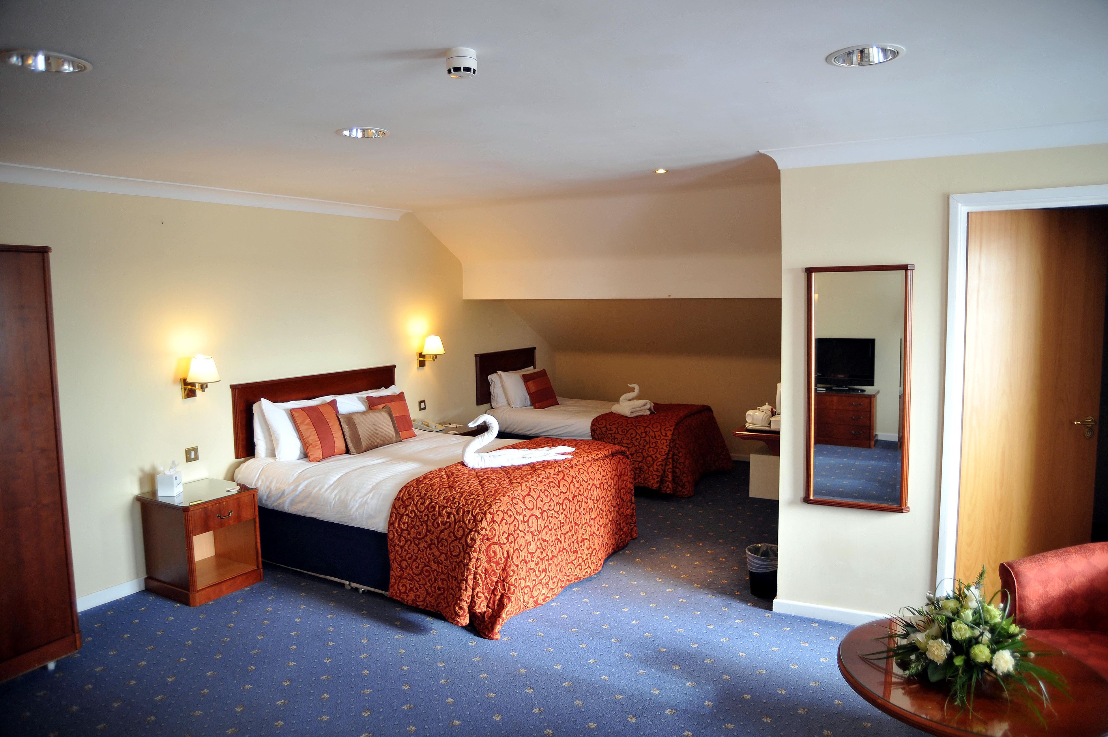 A hotel room with two beds, a small table with a vase of flowers, a mirror, and a wooden wardrobe.