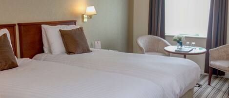 A hotel room with two large beds, a small round table with a vase of flowers, a chair, and a lamp on the wall.