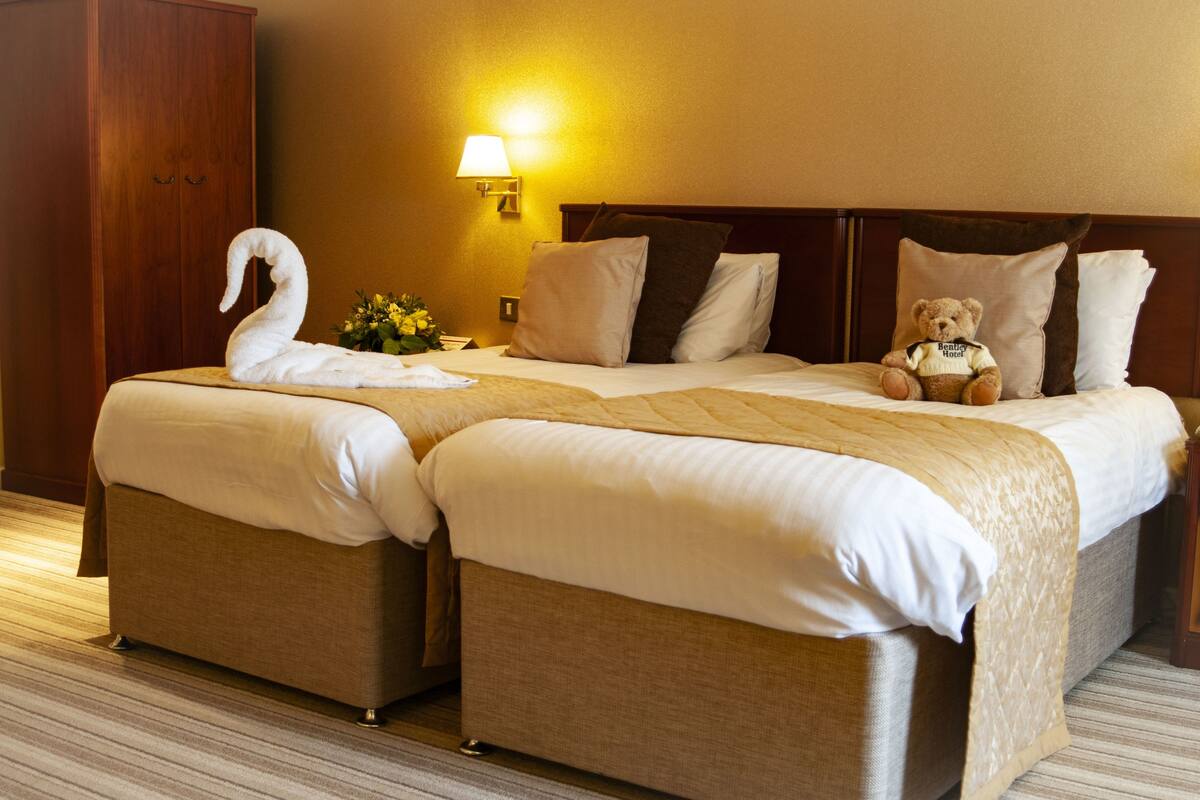 A hotel room with two beds, a wooden wardrobe, a lamp, and a teddy bear on one bed.