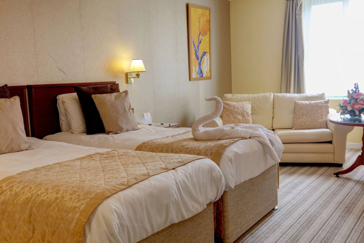 A hotel room with two beds, a sofa, a small table with a vase of flowers, and a painting on the wall.