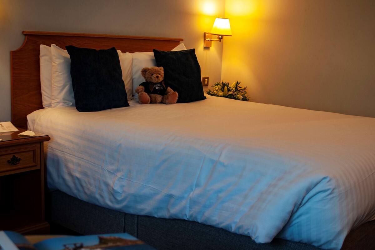 A neatly made bed with white linens, a wooden headboard, and a teddy bear on the pillows.
