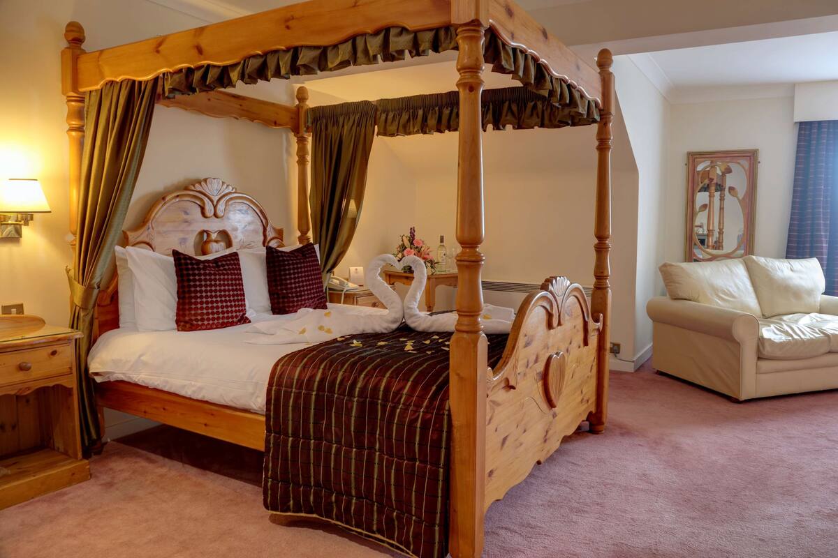 A four-poster bed with a wooden frame and draped curtains, a bedside table with a lamp, and a beige sofa in a room with a carpeted floor.