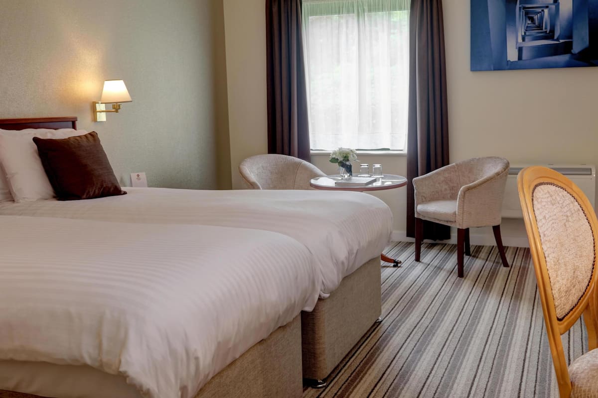 A hotel room with a large bed, two chairs, a small table with a vase, and a painting on the wall.