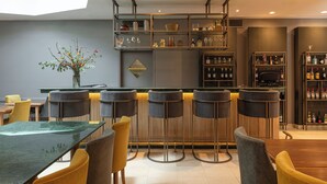Zona de bar moderna con techo alto, taburetes, barra central y estantes con botellas y vasos.