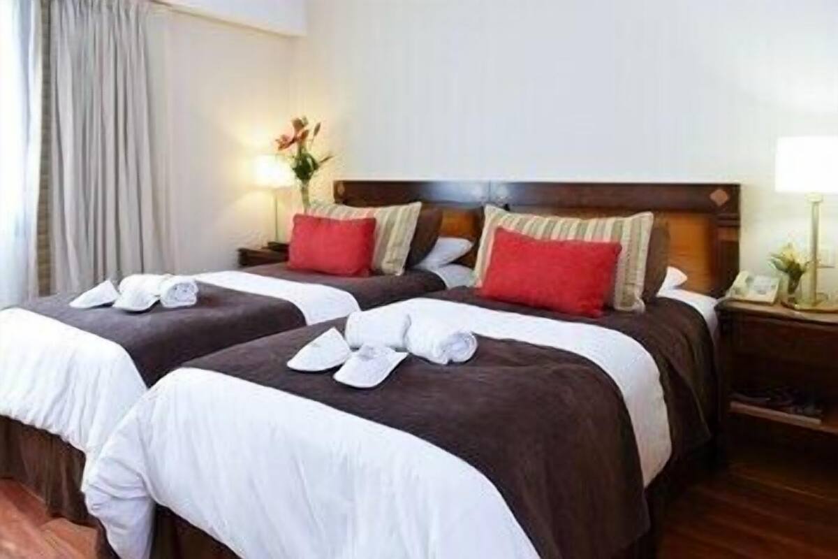 Habitación de hotel con dos camas, cada una con ropa de cama blanca y marrón, almohadas rojas y una mesita de noche pequeña con una lámpara y un jarrón de flores.