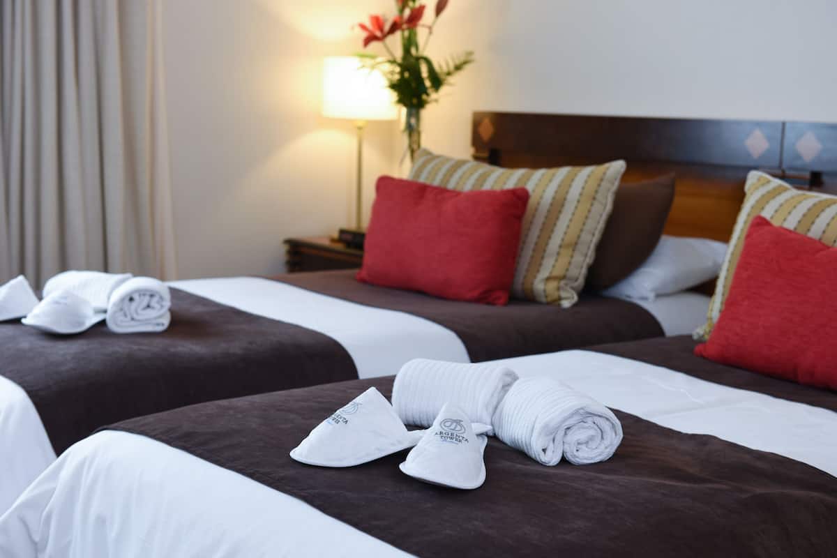 Dos camas con ropa de cama marrón y blanca, almohadas rojas y a rayas, y toallas blancas enrolladas con un logotipo.