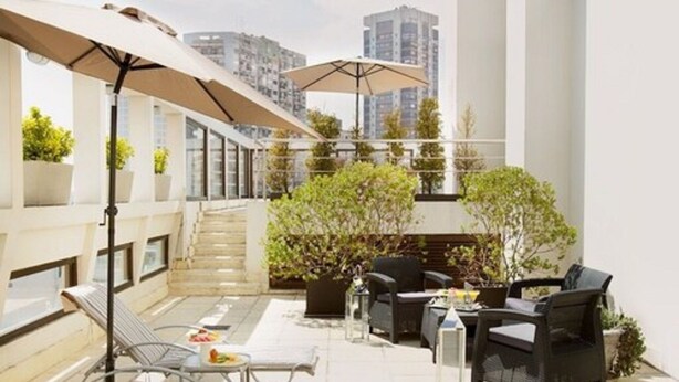 Una terraza en la azotea con mesas al aire libre, plantas en macetas y una escalera que lleva a un nivel superior.