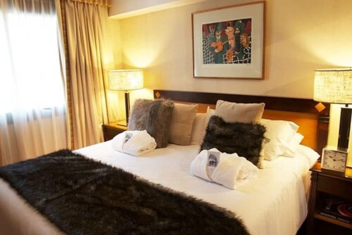 Habitación de hotel con una cama bien hecha, dos lámparas de noche, un cuadro en la pared y una ventana con cortinas.