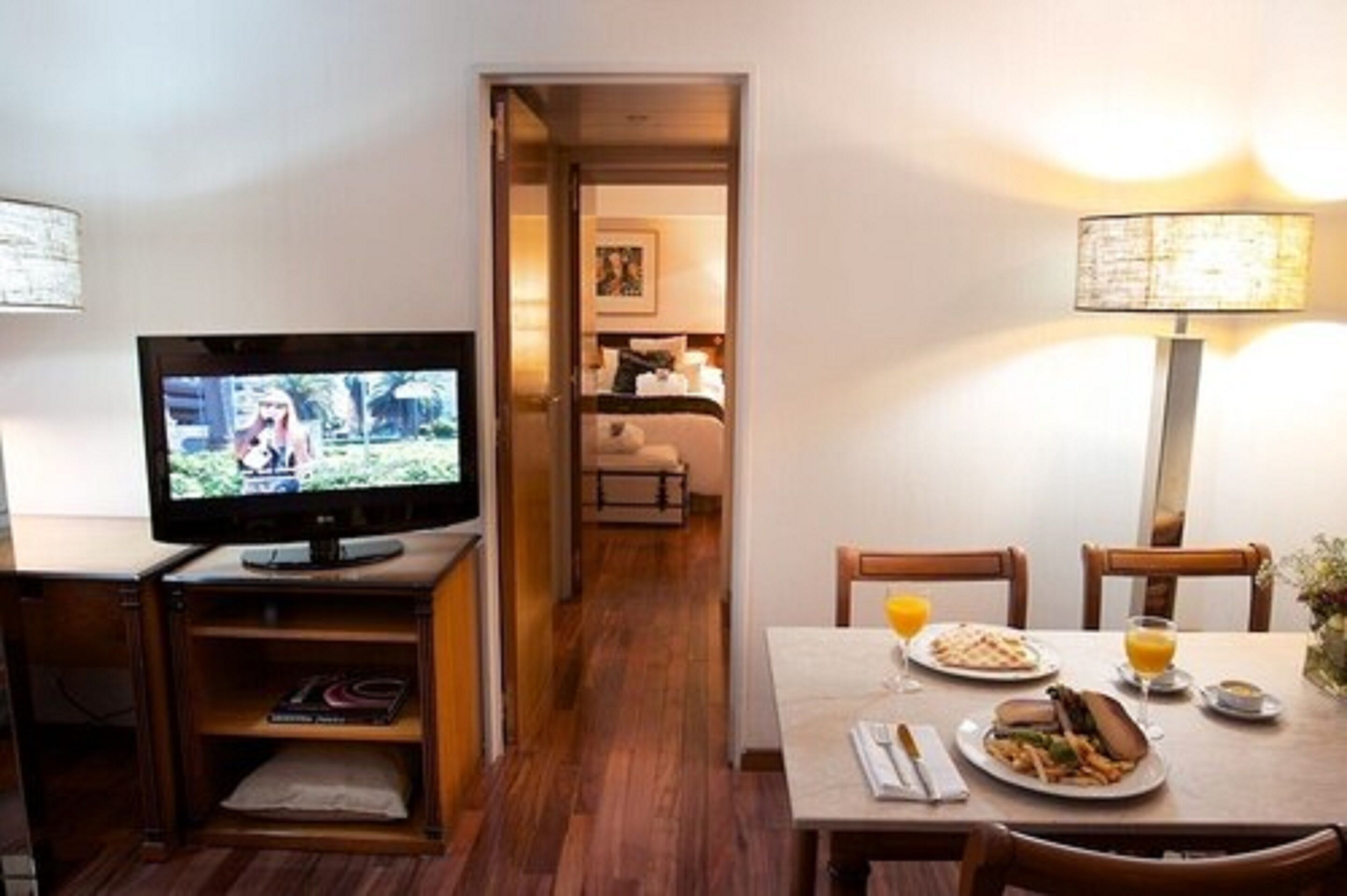 A hotel room with a TV, wooden furniture, a dining table with breakfast, and a hallway leading to a bedroom.
