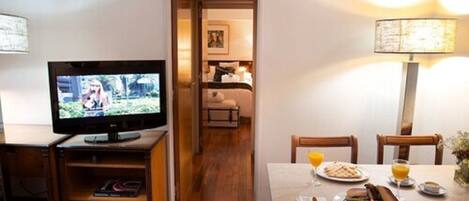 A hotel room with a TV, wooden furniture, a dining table with breakfast, and a hallway leading to a bedroom.