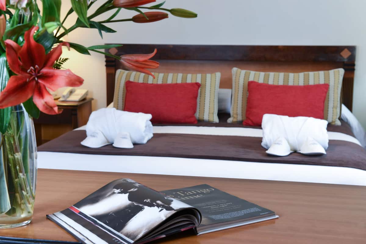 Una cama bien hecha con almohadas rojas y a rayas, un par de toallas blancas y una revista sobre una mesa de madera.