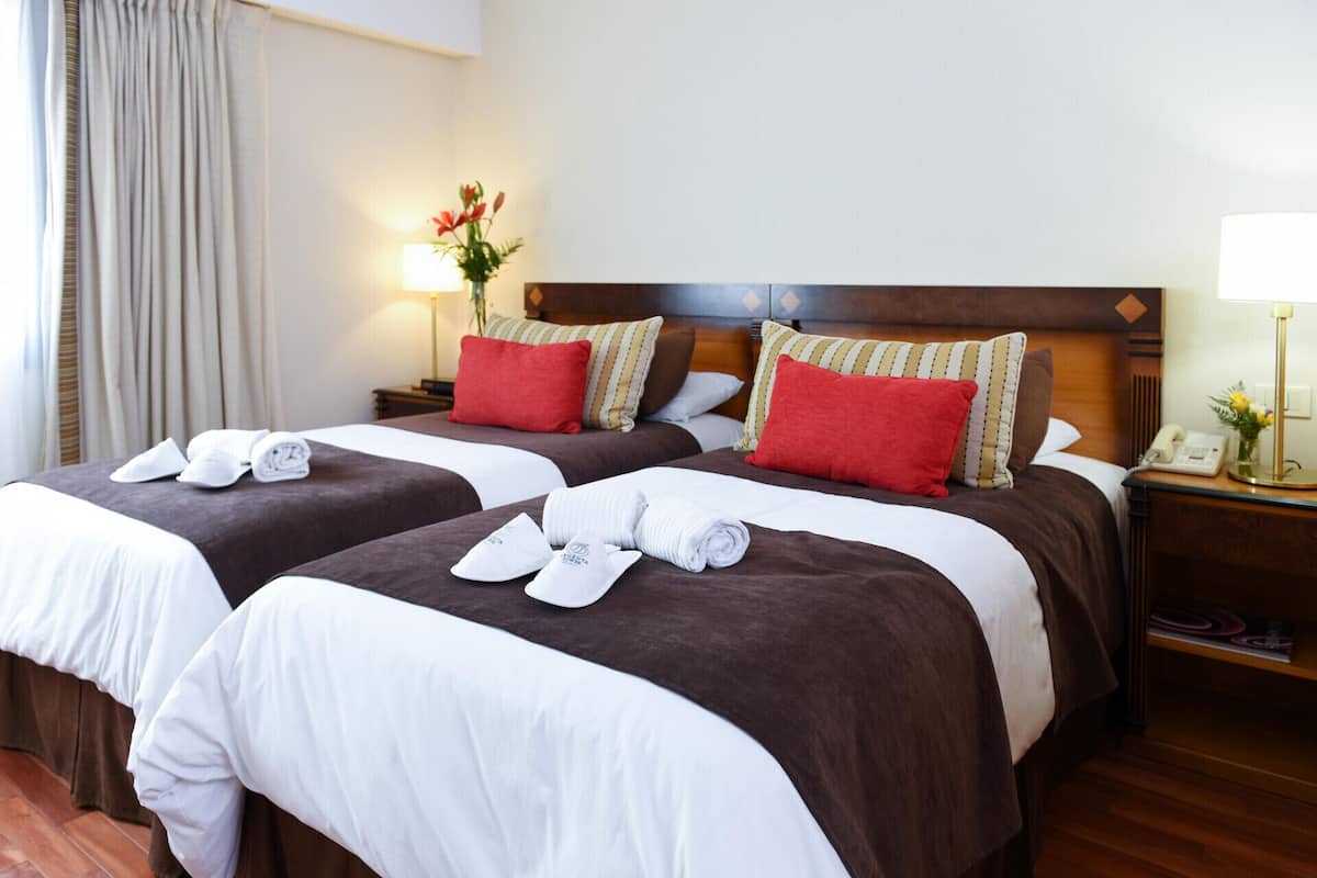 Dos camas con ropa de cama marrón y blanca, almohadas rojas, una mesita de noche con una lámpara, y un florero con flores.