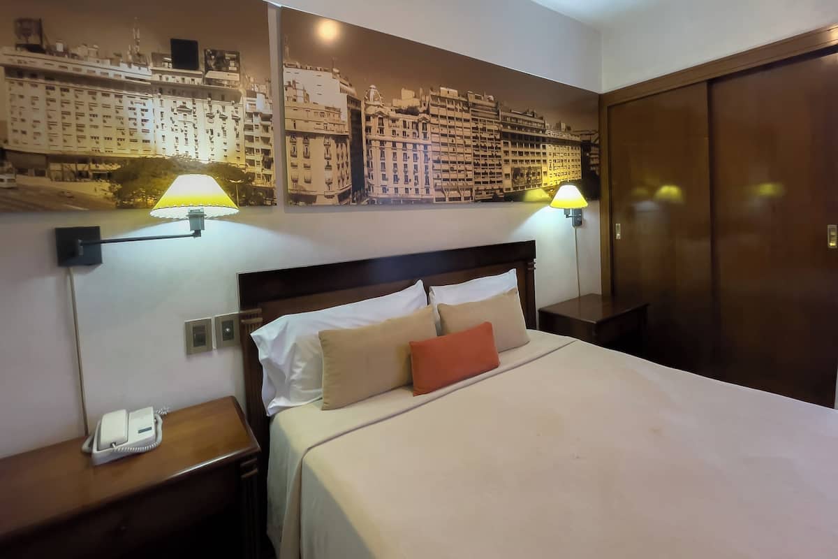A hotel room with a bed, nightstand, wall-mounted lamps, and a cityscape mural.