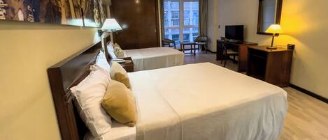 A hotel room with two beds, a desk, a chair, a TV, and a window with a view of buildings.