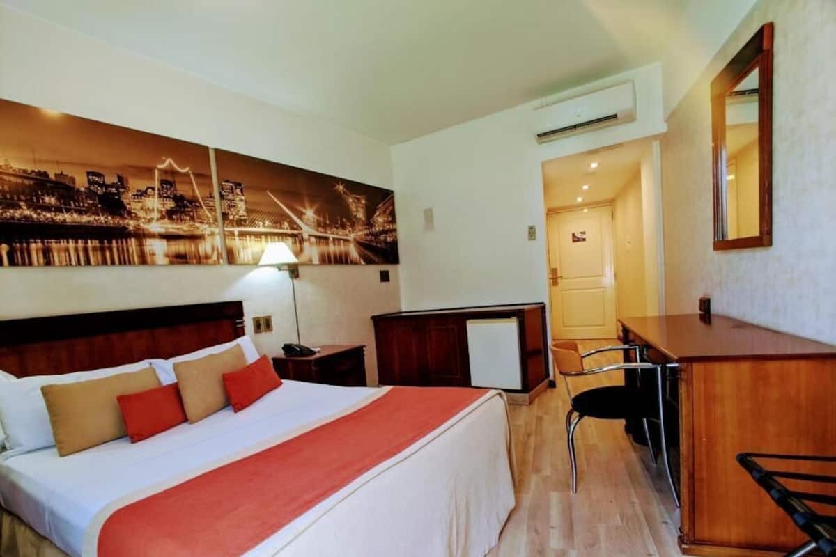 A hotel room with a bed, a desk, a chair, a lamp, and a cityscape painting on the wall.