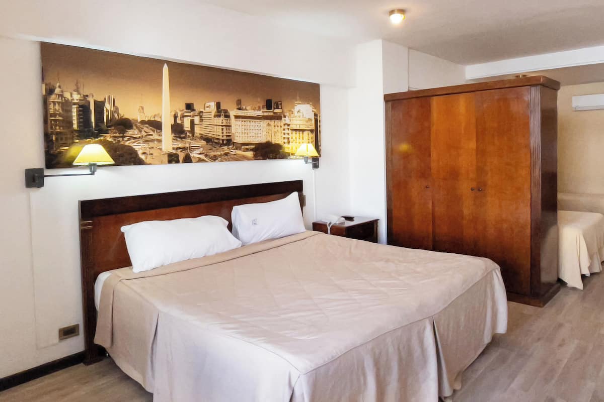 A hotel room with a large bed, a wooden wardrobe, a nightstand, and a cityscape painting on the wall.