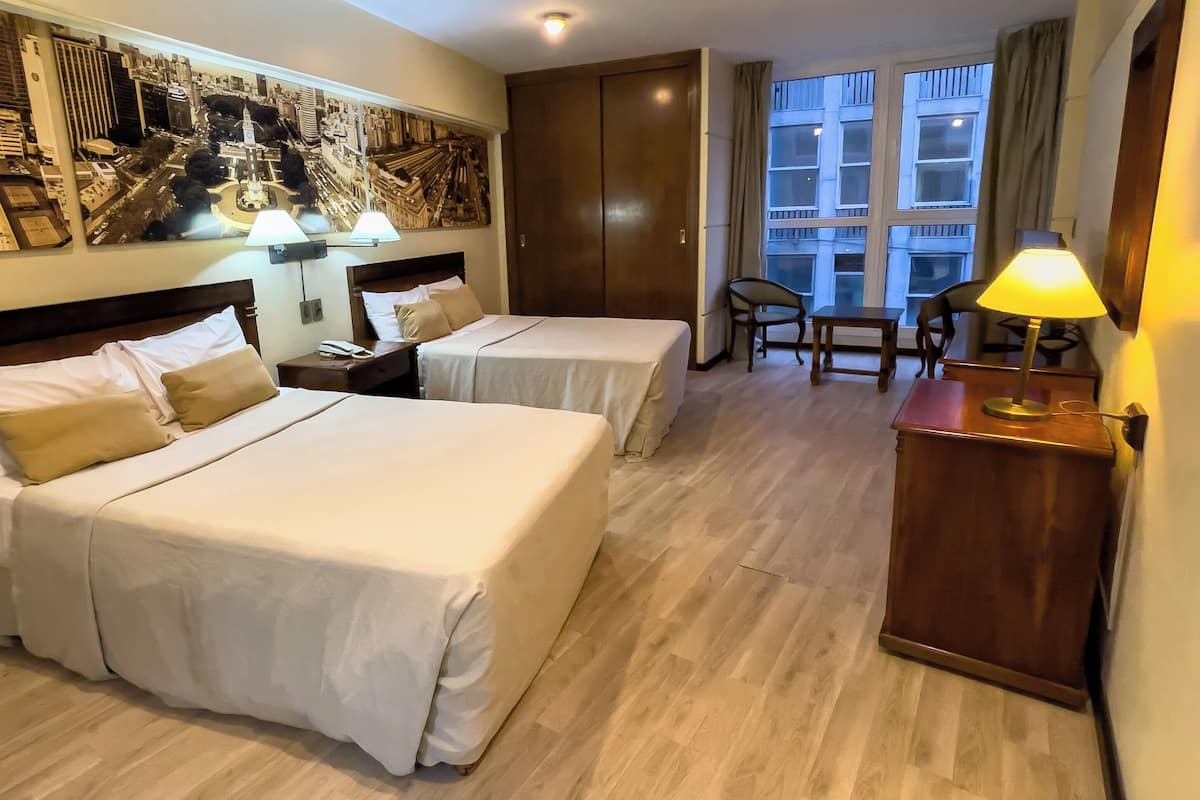 A hotel room with two beds, a desk, a chair, a lamp, and a large window.